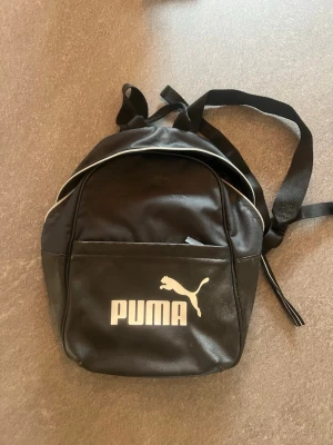 Svart ryggsäck från Puma - Snygg svart ryggsäck från Puma med vit logga framtill och vita detaljer längs dragkedjan. Väskan har ett huvudfack och en ytterficka, justerbara axelband och är tillverkad i ett slitstarkt material som känns lite glansigt. Perfekt för skola eller cityhäng.