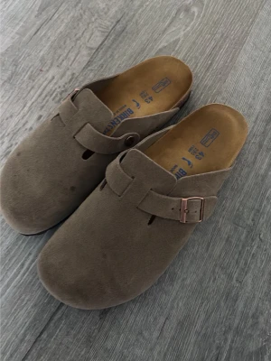 Bruna Birkenstock tofflor mocka - Säljer ett par klassiska bruna Birkenstock tofflor i mocka med justerbart spänne i metall. De har en öppen häl och mjuk innersula för extra komfort. Perfekta för dig som gillar en avslappnad och stilren look.