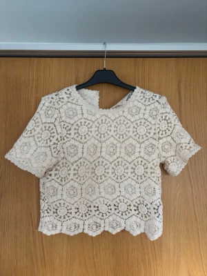 Beige spetsblus med kort ärm - Säljer en superfin beige blus i spets med korta ärmar och rund halsringning. Blusen har ett vackert virkat mönster och knäppning i nacken💕