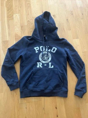 Mörkblå hoodie från Polo Ralph Lauren - Snygg mörkblå hoodie från Polo Ralph Lauren med vit tryckt text och tigerlogga på bröstet. Klassisk känguruficka framtill och justerbar huva med dragsko. Perfekt för en avslappnad och sportig stil.