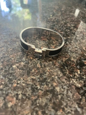 Silverfärgat armband med svart emalj - Snyggt armband i silverfärgad metall med svart emalj och ett H-format spänne framtill. Stelt och stilrent, perfekt för dig som gillar minimalistisk design och vill ha en accessoar som sticker ut lite extra. Omkretsen runt armbandet är 21 cm