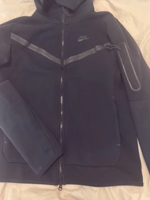 Nike tech fleece - Snygg svart hoodie från Nike med hel dragkedja framtill och huva. Har diskreta reflexdetaljer på bröstet och ärmen samt en liten Nike-logga på vänster sida. Tillverkad i mjukt och stretchigt material, perfekt för en sportig och avslappnad stil.