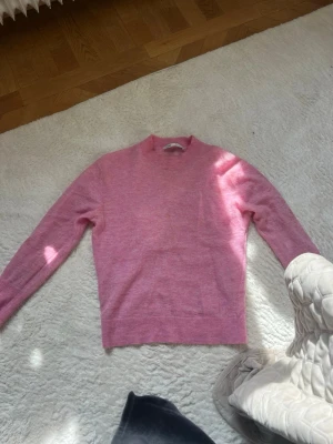 Rosa stickad tröja med rund hals - Säljer en söt rosa stickad tröja med rund hals och långa ärmar. Tröjan har en enkel och clean design, perfekt att matcha med jeans eller kjol. Materialet ser mjukt och skönt ut, perfekt för kyligare dagar.
