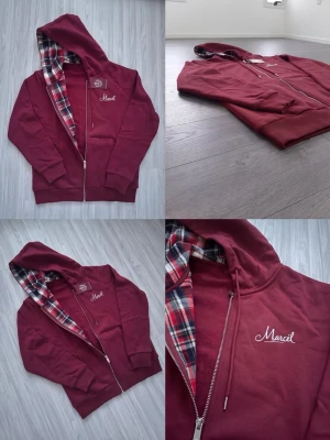 Vinröd hoodie med rutigt foder - Snygg vinröd hoodie med dragkedja och huva. Insidan av huvan har ett rutigt foder i rött, vitt och blått. På bröstet står det 'Marcel' broderat i vitt. Hoodien har fickor framtill och ribbade muddar vid ärmar och nederkant. Perfekt för en chill och avslappnad stil.