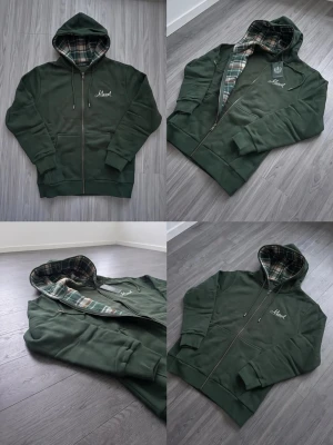 Mörkgrön hoodie med rutigt foder - Snygg mörkgrön hoodie med dragkedja och huva. Insidan av huvan har ett rutigt foder i grönt, vitt och beige. På bröstet finns en liten vit brodyr med texten 'Marvel'. Hoodien har ribbade muddar och fickor framtill.