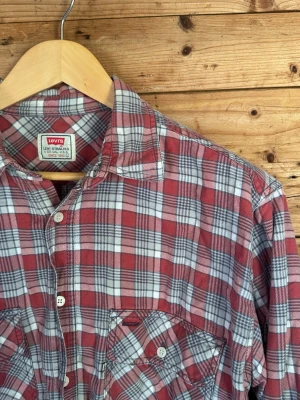 Vintage Levi’s 90s flanellskjorta rutig L - Vintage Levi’s flanellskjorta i röd/grå/vit ruta, strl L. Rejäl 100% bomull med mjuk känsla, två stora bröstfickor och avslappnad boxy passform. Tydlig 90-talslook med naturlig blekning och fin vintagepatina. Fint bruksskick.  Mått: bröst ca 56 cm, axel ca 48 cm, rygglängd ca 73 cm.