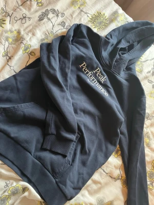 Mörkblå hoodie Peak Performance - Snygg mörkblå hoodie från Peak Performance med broderad logga på bröstet och texten 'Est. 1986'. Klassisk känguruficka framtill, ribbade muddar och skön huva. Perfekt för chill dagar eller när du vill ha en clean look.