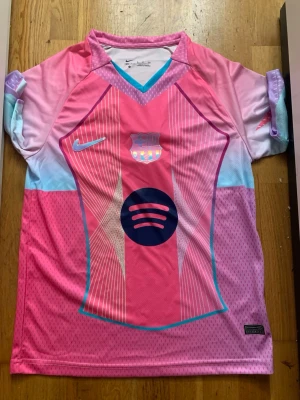 FC Barcelona rosa matchtröja Nike - Snygg FC Barcelona matchtröja från Nike i rosa, lila och ljusblå toner. Tröjan har korta ärmar, klubbmärke och Nike-logga på bröstet samt Spotify-logga i mitten. Tillverkad i lätt och ventilerande funktionsmaterial, perfekt för fotboll eller träning.