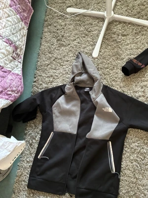 Svart och grå hoodie från The North Face - Säljer en snygg svart och grå hoodie från The North Face med dragkedja framtill och två fickor med dragkedja. Hoodien har justerbar huva med snören och logga på bröstet. Perfekt för chill dagar eller när du vill ha en sportig look.