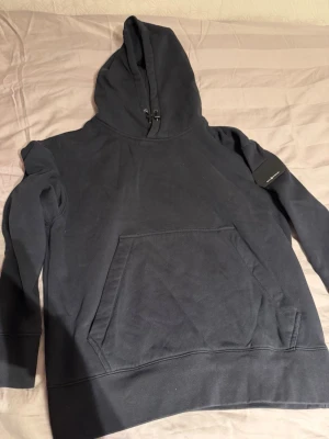 Svart hoodie från Sail Racing - Säljer en svart hoodie från Sail Racing med klassisk känguruficka framtill och justerbar huva. På vänster ärm sitter en svart patch med Sail Racing-loggan. Hoodien är i mjukt bomullsmaterial och har ribbade muddar vid ärmslut och nederkant.