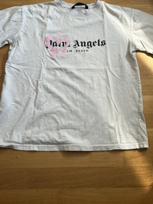 Vit Palm Angels t-shirt med tryck - Säljer en vit t-shirt från Palm Angels med svart text och ett rosa hjärta med drip-effekt på bröstet. T-shirten har rund hals och korta ärmar, perfekt för en avslappnad streetwear-look. Materialet känns mjukt och skönt, troligen bomull.