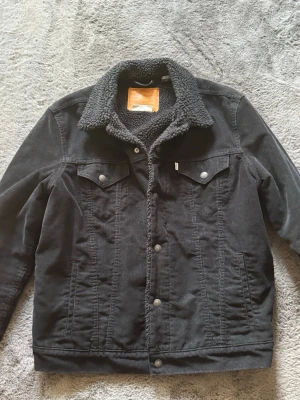 Svart Levis Manchester jacka - Snygg svart jeansjacka med mjuk teddykrage och knäppning framtill. Två bröstfickor med lock och knappar samt sidofickor. Klassisk modell med raka linjer och detaljer i metall. Perfekt för dig som vill ha en stilren och trendig look. L passar M