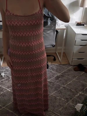 Lång zigzag-mönstrad klänning - Säljer en lång klänning med smala axelband och zigzag-mönster i rosa, beige och brunt. Klänningen har en rak passform och är tillverkad i ett lätt material, perfekt för varma dagar. Snygg och trendig look med boho-vibbar. Storleken är L men för mig som är 165 och väger ca 58 kg sitter den som M