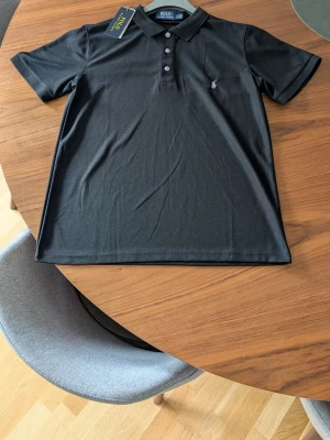 Svart pikétröja från Polo Ralph Lauren - Klassisk svart pikétröja från Polo Ralph Lauren med krage, tre knappar och den ikoniska broderade loggan på bröstet.