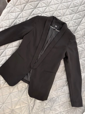Svart kavaj från H&M - Stilren svart kavaj från H&M med klassisk passform och smala slag. Kavajen har två fickor framtill och är helfodrad för en snygg siluett. Perfekt att matcha med skjorta eller t-shirt för en clean look.