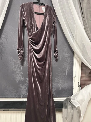 Lång sammetsklänning från NLY Eve - Elegant långklänning i mörk lila sammet från NLY Eve. Klänningen har djup v-ringning, långa ärmar och draperad omlottdesign framtill. Perfekt för dig som vill sticka ut med en lyxig och trendig look.
