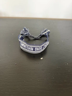 Blått Christian Dior tygarmband - Snyggt blått tygarmband från Christian Dior med texten 'J'ADIOR' invävd i vitt. Armbandet har justerbar knytning med tofsar i ändarna och en cool, bohemisk känsla. Perfekt accessoar för att lyfta din outfit.