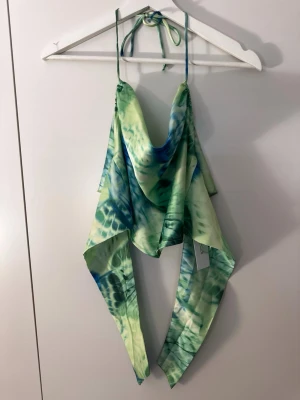 Grön och blå omlott linnetopp - Trendig omlott linnetopp i grönt och blått med knytband i nacken. Toppen har ett unikt mönster och tunna axelband som ger en cool och somrig vibe. Perfekt för dig som vill sticka ut med en färgstark och luftig topp.