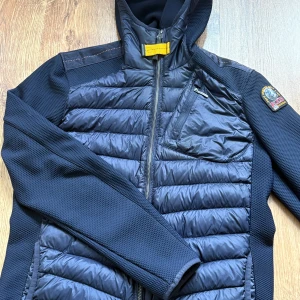 Svart hybrid jacka  från Parajumpers - 1699kr nypris 3899. Snygg svart hybridjacka från Parajumpers med huva och gult spänne vid halsen. Jackan har quiltad framsida och rygg, dragkedja, bröstficka med zip och patch på ärmen. Materialmix av polyester och syntet ger både värme och sportig känsla. Använd några gånger inget som syns dock 