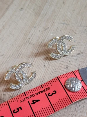 Örhängen med strass i nyskick  - Säljer ett par ikoniska örhängen i guldtonad metall med glittrande strass på de klassiska dubbel-C-loggorna. Stift med transparent bakstycke. Perfekta för dig som vill ha en lyxig och trendig detalj till din look.