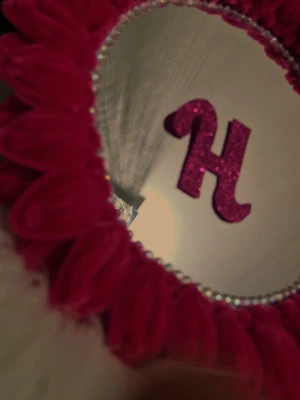 Rund spegel med rosa dekoration - Unik rund spegel med ram av rosa konstblommor och pärlband runt spegelglaset. Spegeln har en personlig touch med ett glittrigt H i mitten men initialen kan ändras och anpassas efter vad du önskar. Perfekt som dekorativ detalj på sminkbordet eller väggen.
