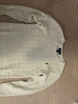 Vit kabelstickad tröja Polo Ralph Lauren - Kabelstickad vit tröja från Polo Ralph Lauren med rund halsringning och klassisk marinblå logga broderad på bröstet. Tröjan har långa ärmar och ribbade muddar vid ärmslut och nederkant. Tillverkad i mjukt material som känns lyxigt. Har en liten defekt på frmasidan där jag sytt men man inte tänker på när man har på sig den.