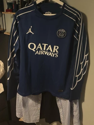 Dembele Psg longsleeve - Säljer en blå långärmad PSG matchtröja med vita detaljer och vingmönster på ärmarna. Framsidan har Qatar Airways-logga och Paris Saint-Germain-märke, baksidan har O. Dembélé och nummer 10 tryckt. Perfekt för fotbollsfans!
