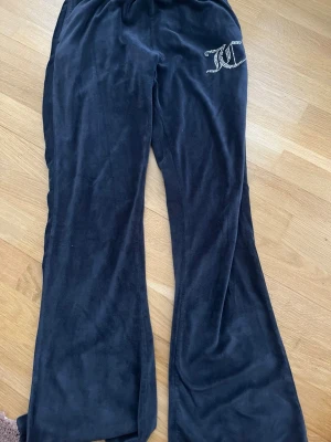 Svarta mjukisbyxor Juicy Couture - Mysiga svarta mjukisbyxor från Juicy Couture i velour med glittrande JC-logga på höften. Byxorna har utsvängda ben och resår i midjan för extra komfort. Perfekta för en chill och trendig look. Fickorna kan vara isönder men går att sy ihop