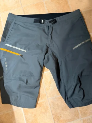 Mörkgrå funktionsshorts med snörning - Snygga mörkgrå funktionsshorts med justerbar snörning i midjan och flera fickor, inklusive en med orange dragkedja. Tillverkade i ett slitstarkt syntetmaterial, perfekta för aktiva dagar utomhus. Reflekterande detaljer för extra synlighet.