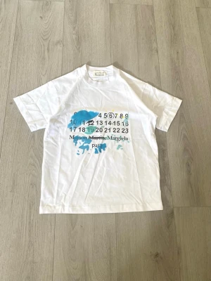 Maison Margiela vit t-shirt med tryck - Säljer en vit t-shirt från Maison Margiela med coolt tryck framtill i blått och svart, samt siffror och logga. T-shirten har rund hals och korta ärmar. Perfekt för dig som gillar streetwear och unika designer.