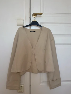 Beige vårjacka med vida ärmar - Stilren beige vårjacka med croppad passform och vida ärmar. Koftan har dekorativa sömmar längs kanterna och breda muddar vid ärmsluten. Perfekt att slänga över en topp för en avslappnad look. ( pris kan diskuteras)