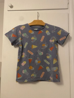 Blå t-shirt med glassmönster Polarn O. Pyret - Säljer en blå t-shirt från Polarn O. Pyret med färgglatt glassmönster i olika pastellfärger. T-shirten har rund halsringning och korta ärmar. Perfekt för dig som gillar lekfulla prints och vill sticka ut lite extra i vardagen.