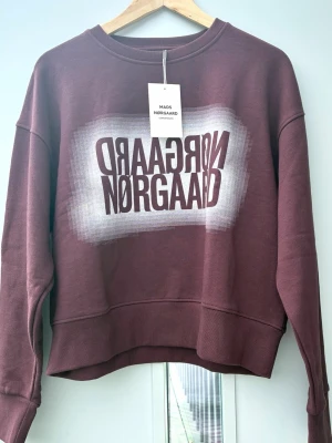 Vinröd sweatshirt från Mads Nørgaard - Cool vinröd sweatshirt från Mads Nørgaard med tryckt logga i vitt och vinrött på bröstet. Tröjan har rund hals, långa ärmar och ribbade muddar. Tillverkad i mjukt bomullsmaterial för en avslappnad och trendig look.