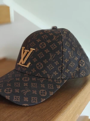 Brun med monogram - Snygg brun keps med monogram i guldbrunt över hela kepsen. Böjd skärm och broderad logga framtill. Tillverkad i ett slitstarkt tygmaterial som ger en exklusiv känsla.