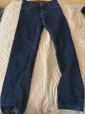 Mörkblå jeans från Hugo - Säljer ett par mörkblå jeans från Hugo med klassisk femficksdesign och raka ben. Jeansen har röda detaljer vid linningen och på fickan, samt tydliga sömmar. Perfekta för en avslappnad och stilren look.