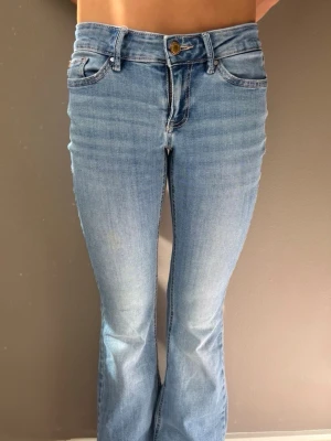 Ljusblå bootcut jeans - Säljer ett par ljusblå bootcut jeans med klassisk femficksdesign och låg midja. Jeansen har en snygg tvätt med lätt slitning framtill och är tillverkade i stretchigt denimtyg för skön passform. Perfekta till sneakers eller boots. De har en liten gul fläck som inte är så synlig när jeansen är på. 