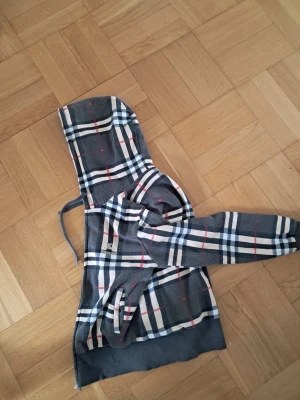 Grå Burberry hoodie med rutigt mönster - Snygg grå hoodie från Burberry med klassiskt rutigt mönster i vitt, svart och rött. Hoodien har huva med dragsko och broderad Burberry-logga på bröstet. Perfekt för dig som vill sticka ut med en ikonisk streetstyle-look.