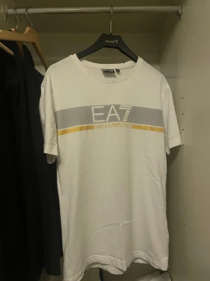 Vit EA7 Emporio Armani t-shirt - Snygg vit t-shirt från EA7 Emporio Armani med tryck i silver och guld på bröstet. Klassisk rund hals och korta ärmar. Perfekt till jeans eller shorts för en clean och stilren look. 