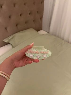 Grön hårklämma med text och hjärtan - Söt hårklämma i ljusgrönt med texten 'Matcha mood' i rosa och fyra glittriga hjärtan framtill. Klämman har en vågig form och är gjord i plast, perfekt för att piffa till frisyren med en trendig detalj.