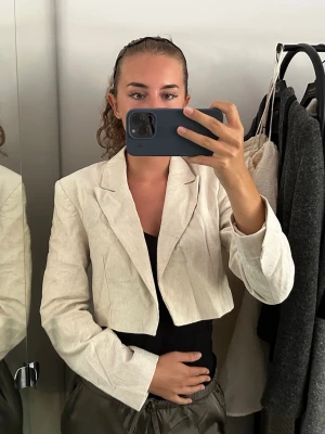 Beige croppad kavaj i linne - Trendig croppad kavaj i beige linne-look med klassisk krage och långa ärmar. Perfekt för dig som vill ha en stilren och modern touch till outfiten. Passar snyggt till både byxor och kjol. Enkelknäppt framtill och lätt oversized känsla.