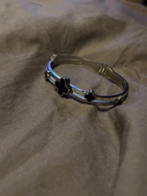 Silverfärgat armband med svarta blommor - Snyggt armband i silverfärg med två svarta fyrklöverformade detaljer och små gnistrande stenar längs bandet. Armbandet har en stilren och modern design som passar perfekt till din outfit. Lätt att ta på tack vare smidig öppning.