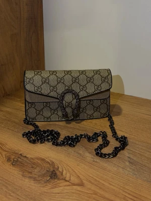 Gg axelremsväska - Axellväska i beige canvas med GG-monogram över hela väskan. Väskan har en metallfärgad kedjerem och ett ikoniskt spänne framtill. Stilren och kompakt modell med svarta kanter och detaljer. Perfekt för dig som gillar lyxiga accessoarer.