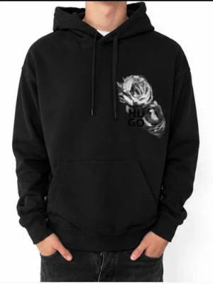 Svart hoodie från Hugo med rosor - Svart hoodie från Hugo med stor huva och dragsko. Framtill finns en vit och grå ros tillsammans med Hugo-loggan, och på ryggen syns flera rosor och stor logga. Klassisk känguruficka och ribbade muddar. Materialet är mjukt bomullstyg. Passar från 165 till 180 cm beroende på vikt samt hur du har dina byxor 