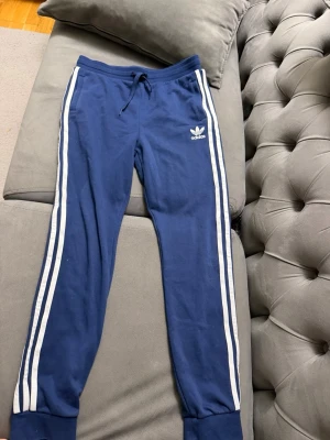 Blå Adidas mjukisbyxor med vita ränder - Snygga blå mjukisbyxor från Adidas med klassiska vita ränder längs benen och dragsko i midjan. Byxorna har muddar vid bensluten och Adidas-logga på vänster lår. Perfekta för chill eller träning.