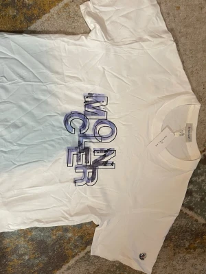 Vit Moncler t-shirt med tryck - Säljer en vit t-shirt från Moncler med stort grafiskt tryck i blå och lila toner på bröstet. Klassisk rund halsringning och korta ärmar. Liten Moncler-logga på ärmen. Perfekt för dig som gillar streetwear och vill sticka ut.