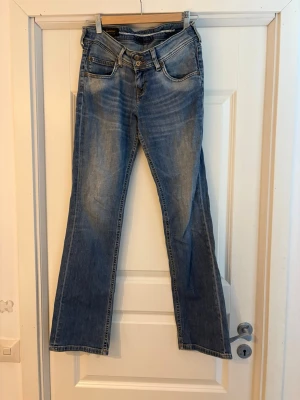 Blåa Lågmidjade bootcut jeans med dubbla knappar - Ett par klassiska blå Lågmidjade  bootcut jeans med dubbla knappar i midjan. Jeansen har en lätt tvättad look och utsvängda ben. Perfekta för dig som gillar retrostil och vill ha ett par bekväma jeans i bomull.