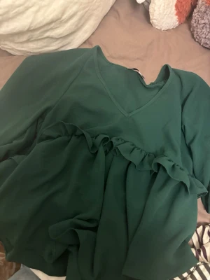 Mörkgrön blus med volang från ASOS - Supersnygg mörkgrön blus från ASOS DESIGN med v-ringning och volangdetalj över bröstet. Blusen har långa ärmar och är gjord i ett mjukt, lätt tyg som faller fint. Perfekt att styla med jeans eller kjol för en trendig look.