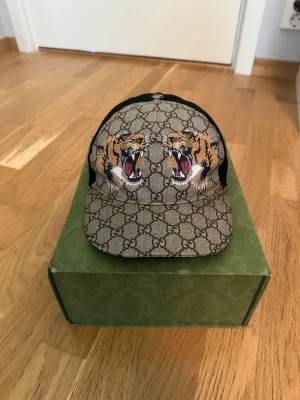 Gucci keps med tigertryck - Tja säljer nu en snygg Gucci keps med klassiskt GG-mönster och två stora tigerhuvuden framtill. Kepsen har justerbart band bak med grön-röd rand och detaljer i svart mesh. Perfekt för dig som vill sticka ut med en exklusiv accessoar.