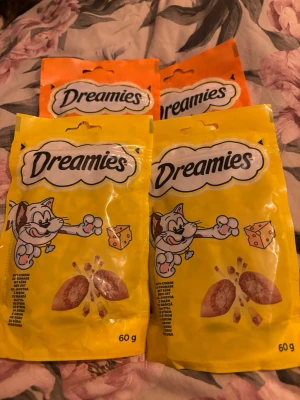 Dreamies kattgodis 4 påsar - Fyra påsar Dreamies kattgodis, två i gul förpackning med ostsmak och två i orange förpackning. Varje påse innehåller 60 gram och har en tecknad katt på framsidan. Perfekt för att skämma bort din katt med något extra gott.