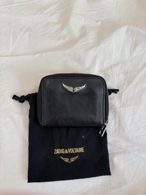 Svart plånbok från Zadig & Voltaire - Snygg svart plånbok i äkta läder från Zadig & Voltaire. Plånboken har en silverfärgad logga med vingar på framsidan och dragkedja runt om. Kommer med original dustbag. Perfekt storlek för kort och kontanter.